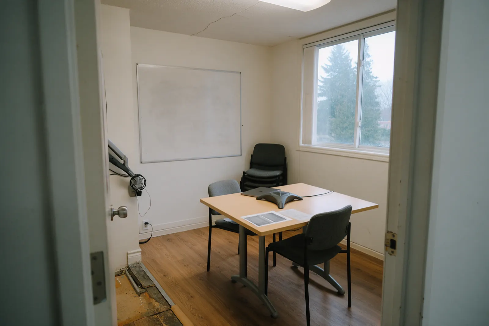 Nothing is in motion. A grey spider conference phone sits in the middle of a light wood table. Two printed one-page sheets lie face-up at a grazing angle so type is grey....